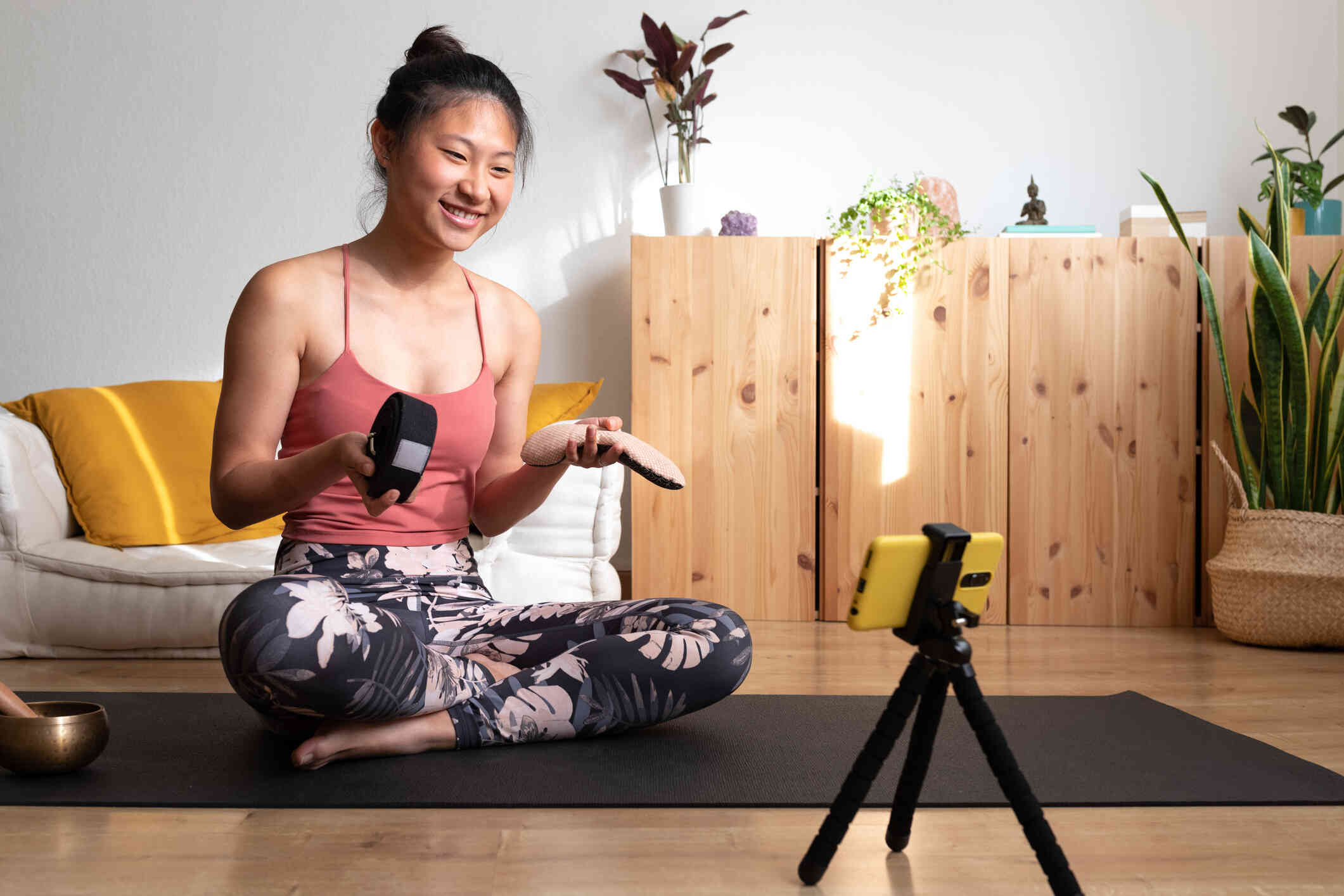 A woman in workout clothes sits on the floor with her phone infront of her on a tripod while smiling as she films a video.